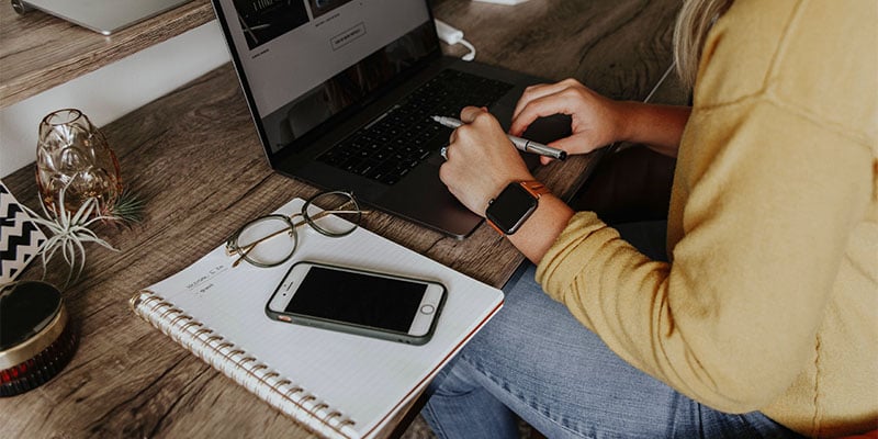 A worker sits in their office space working on their laptop with their phone and notebook beside them.